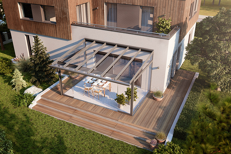 Moderner Wintergarten mit Glasdach und Glaswänden an einem weißen Haus, mit Esstisch und Stühlen innen, umgeben von Holzterrasse und Garten