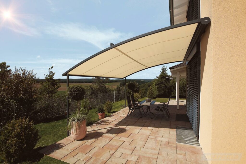 Terrasse mit beige-farbenem gebogenen Sonnensegel, Gartenmöbeln und Pflanzen vor einem Haus mit gelber Fassade