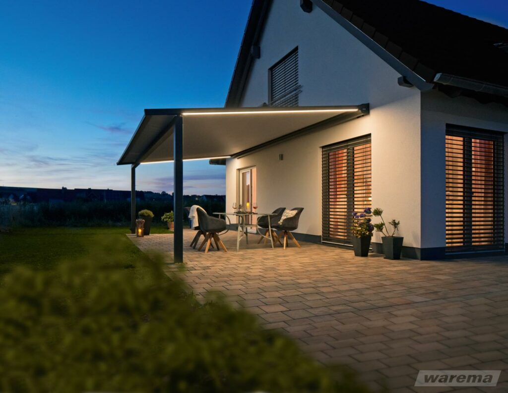 Terrasse eines modernen Hauses bei Dämmerung mit beleuchtetem, ausfahrbarem Sonnenschutz und Sitzgruppe mit Tisch und Stühlen