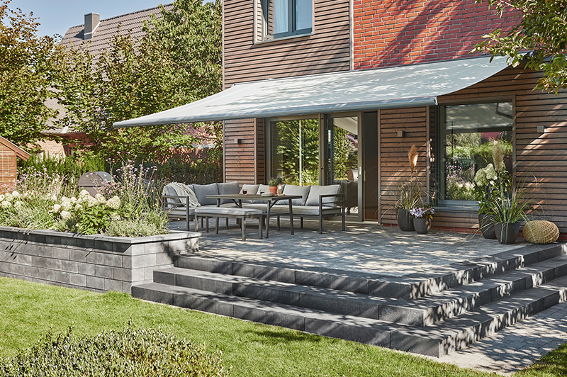 Terrasse mit grauem Sitzmöbel-Set und ausfahrbarer weißer Markise an einem Haus mit Holz- und Ziegelfassade, umgeben von Garten mit Blumen und Rasen