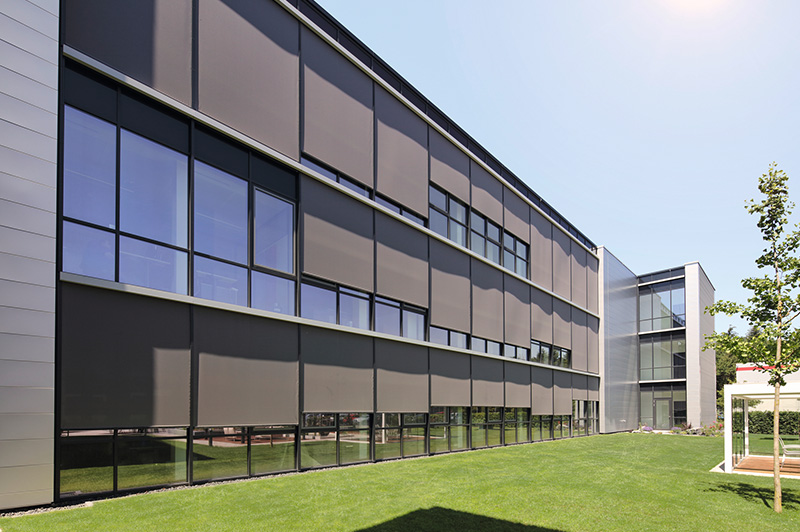 Modernes Bürogebäude mit großen Fenstern, die teilweise mit braunen Sonnenschutzrollos bedeckt sind, umgeben von grünem Rasen und einzelnen Bäumen