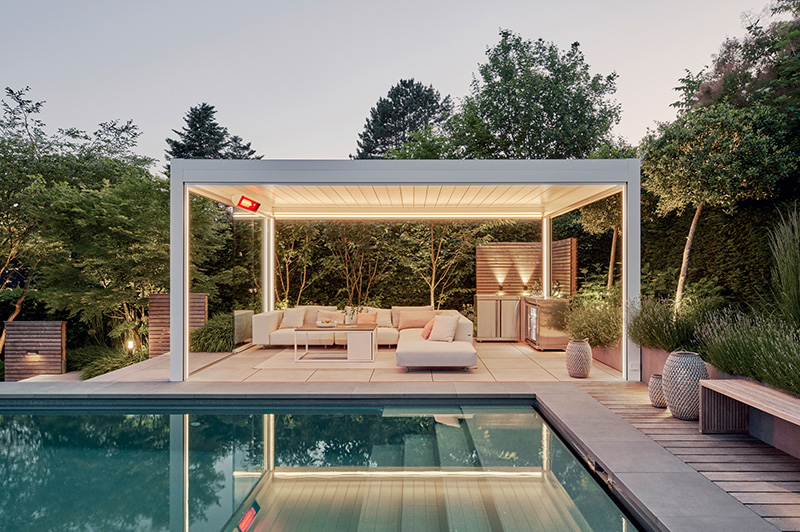 Moderne, beleuchtete Überdachung mit Sitzbereich und Sofas neben einem Pool, umgeben von Garten und Bäumen
