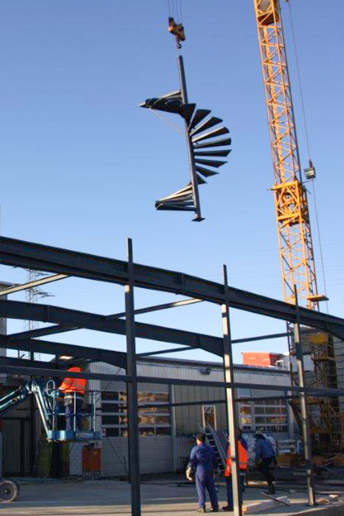 Ein Kran der mit einer Metalltreppe beladen ist und diese an die richtige Position bringt. Monteure die unten am Boden stehen geben Anweisungen und warten auf die Treppe