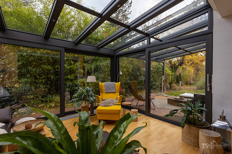 Wintergarten mit Aluminiumrahmen, Glasdach und großen Fenstern, gelber Sessel, Pflanzen und Blick auf herbstlichen Garten