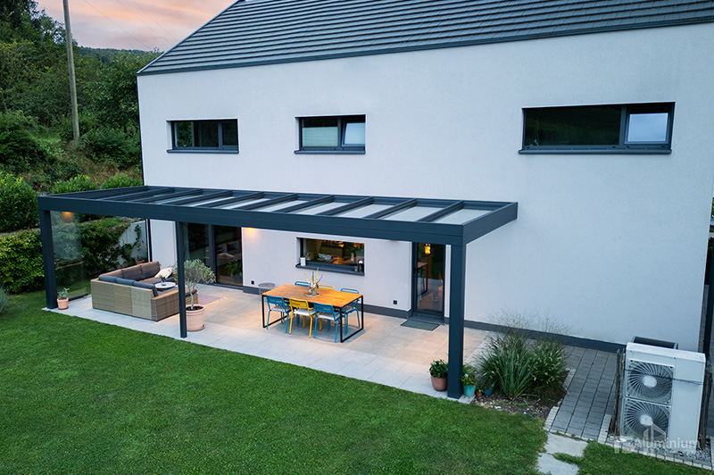 Moderner weißer zweistöckiger Haus mit einer Aluminiumüberdachung über der Terrasse, darunter ein Esstisch mit Stühlen und eine Sitzecke mit Sofa