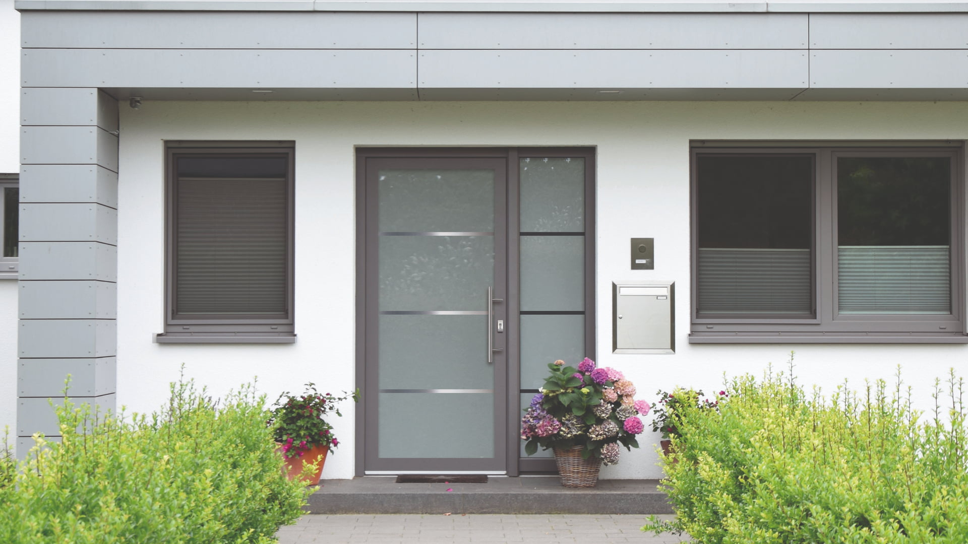 Eingang eines modernen Hauses mit grauer Tür, zwei Fenstern mit geschlossenen Rollläden und Blumenkörben links und rechts der Tür