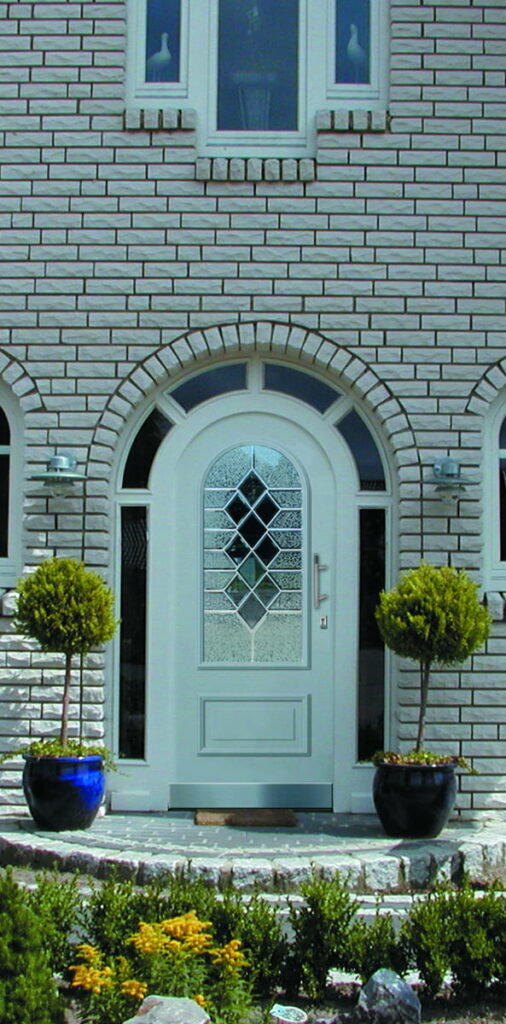 Eingangstür mit Bogenfenster und dekorativem Glas, umgeben von weißem Mauerwerk und zwei kleinen Bäumen in blauen Töpfen