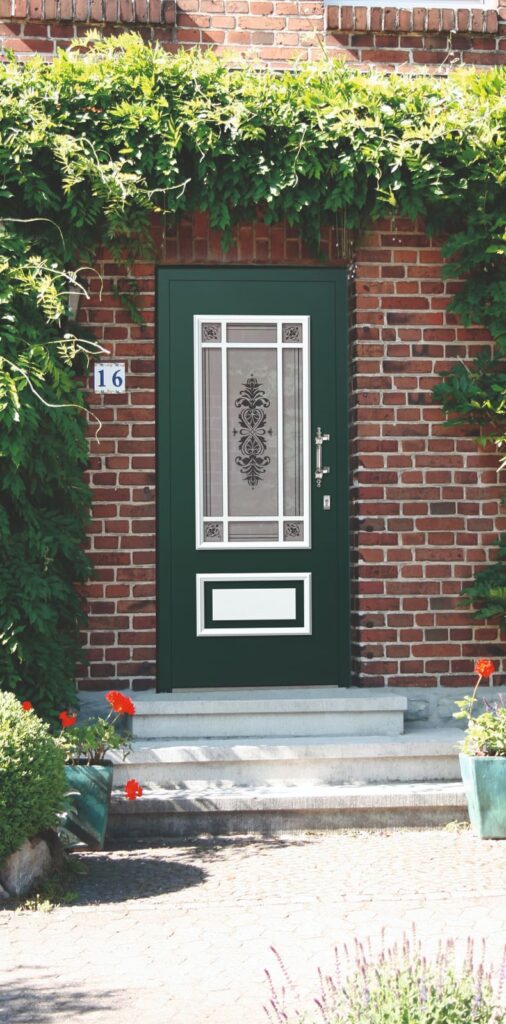 Grüne Haustür mit dekorativem Glasfenster in einem roten Backsteinhaus, umgeben von grünen Pflanzen und Blumen