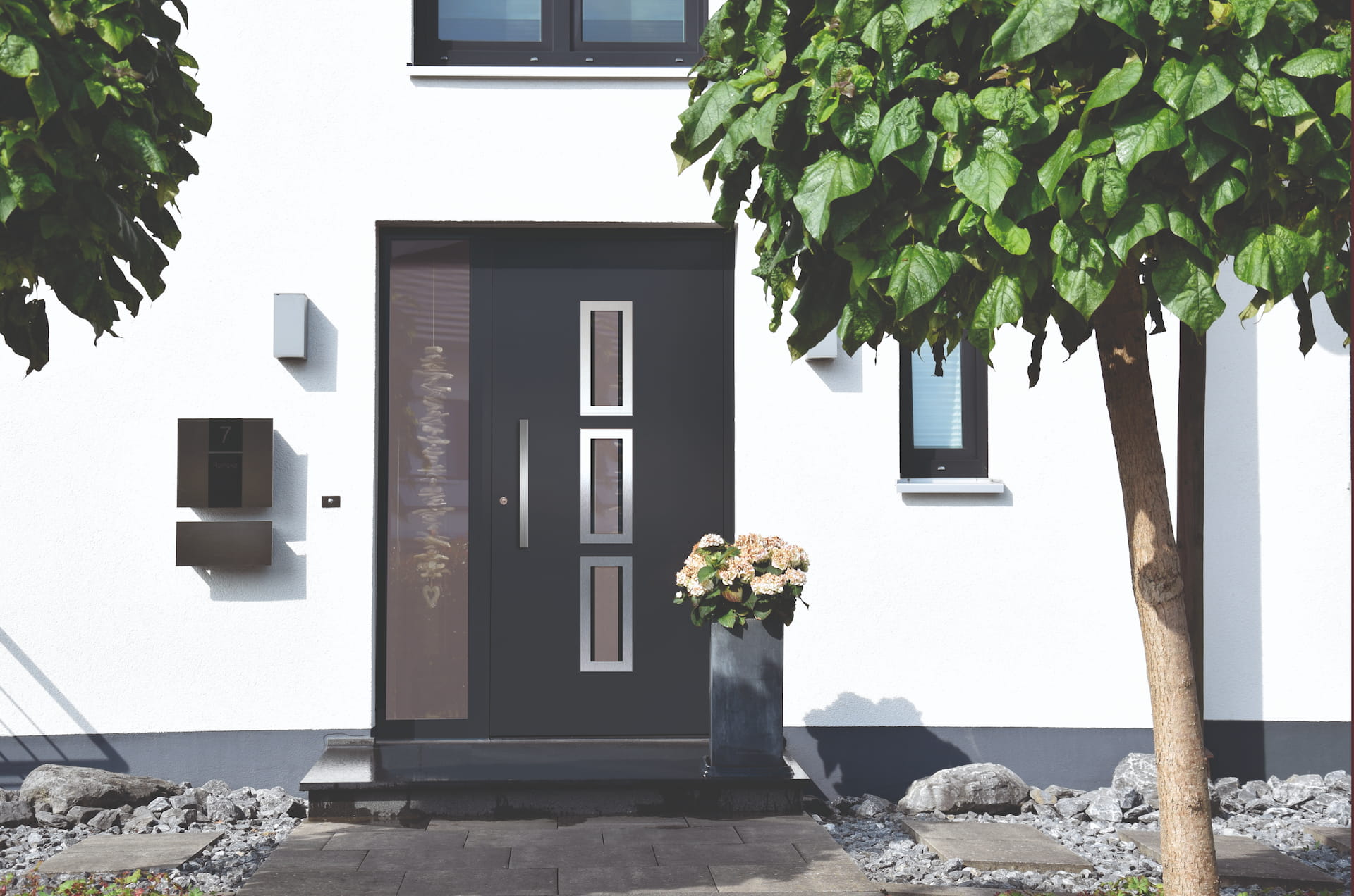 Moderner Hauseingang mit schwarzer Tür, drei rechteckigen Fenstern und Blumendekoration neben dem Eingang