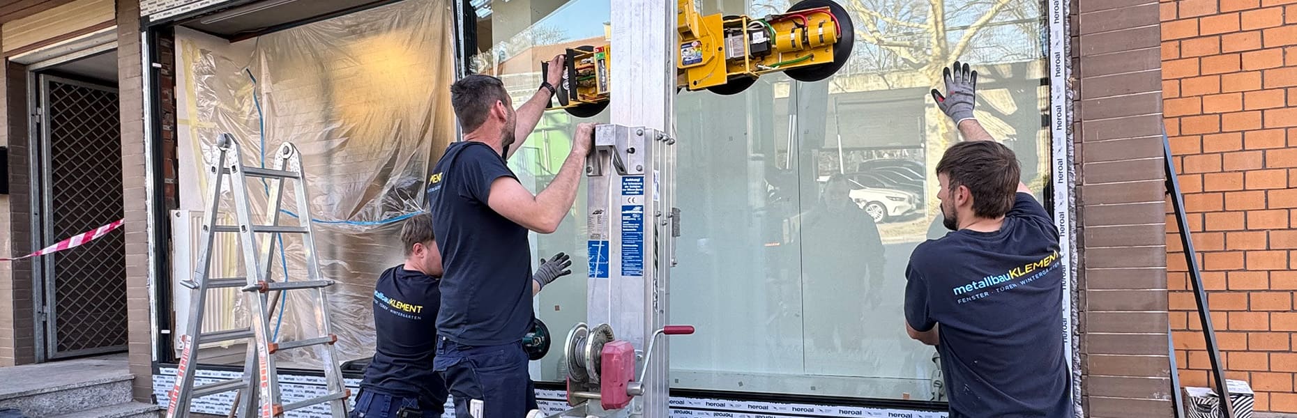 Drei Handwerker montieren eine große Glasscheibe an einem Gebäude, verwenden Hebewerkzeuge und tragen dunkelblaue T-Shirts mit dem Firmenlogo