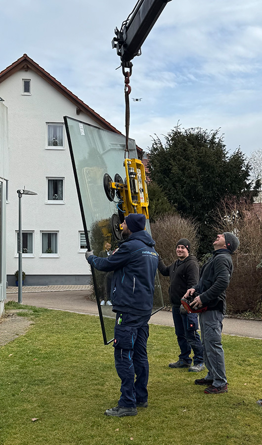 Drei Arbeiter heben mit einem Kran ein großes Glasfenster vor einem Wohnhaus