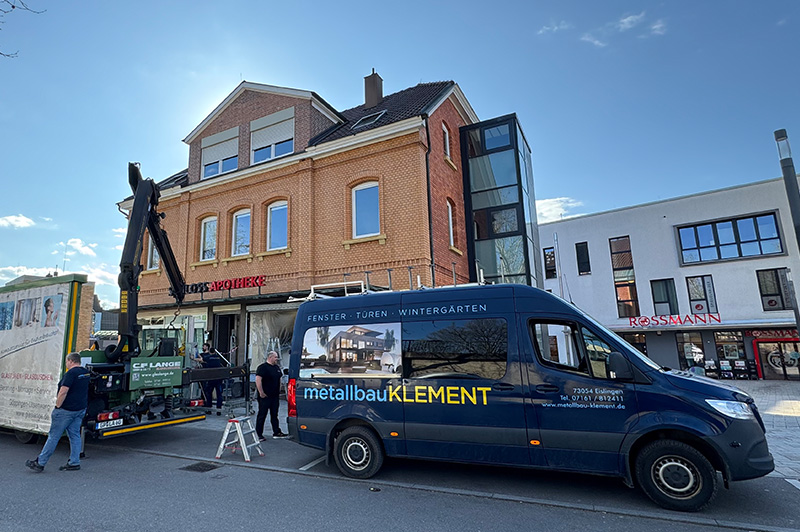 Metallbau Klement Transporter vor einem roten Backsteingebäude mit Aufschrift 'Apotheke', Arbeiter bedienen Kran an einem LKW