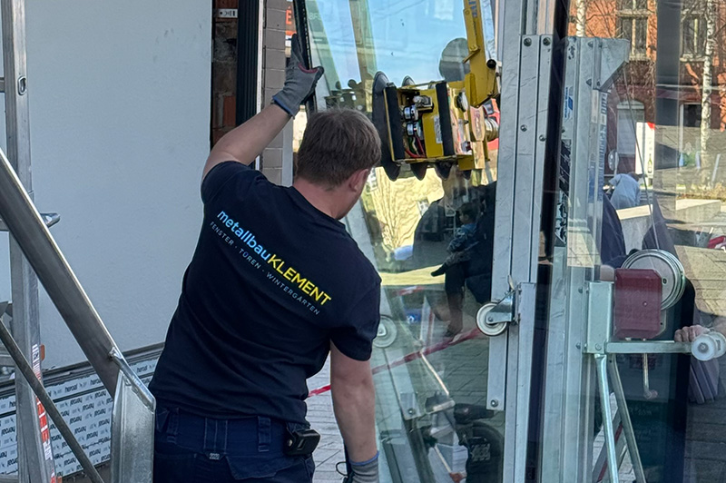 Handwerker mit dunkelblauen T-Shirt und Handschuhen montiert große Glasscheibe mit Saugnapfheber an einem Gebäude
