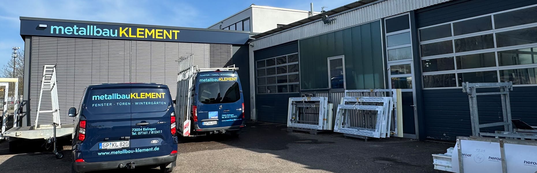 Außenansicht der Werkstatt von Metallbau Klement mit zwei blauen Firmenfahrzeugen und gelagerten Fensterrahmen
