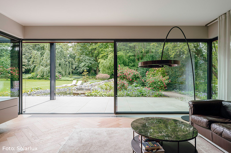 Wohnzimmer mit großen bodentiefen Fenstern von Solarlux, Blick auf Garten mit Rasen, Teich und blühenden Sträuchern