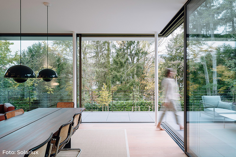 Moderner Essbereich mit großem Holztisch und schwarzen Stühlen, angrenzend an eine Terrasse mit Glas-Schiebetüren und Blick auf herbstlichen Wald